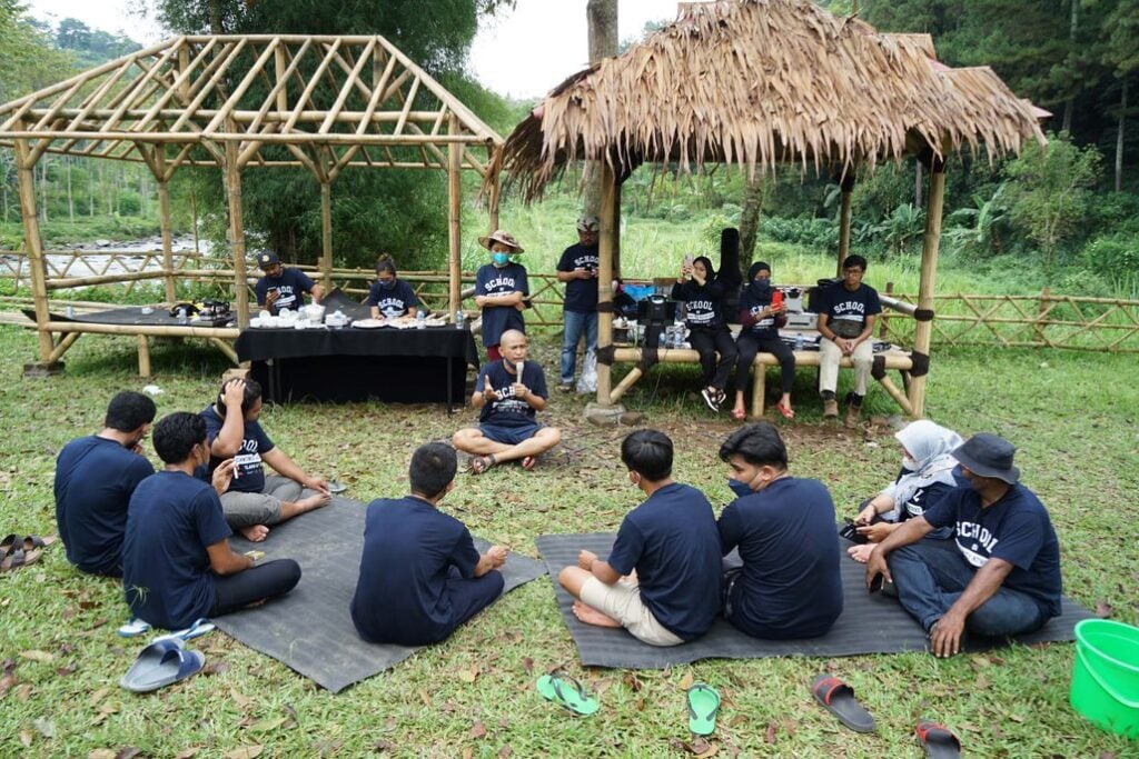 A group of students sit on mats on the ground outside while a teacher sits and speaks at a microphone. There are also bamboo structures providing shade for some students.
