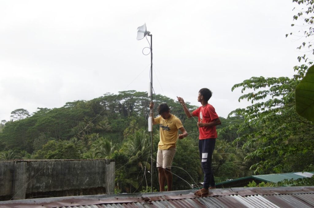 Two people on a roof, surrounded by forest, setting up a P2P connection in San Isidro, Philippines.