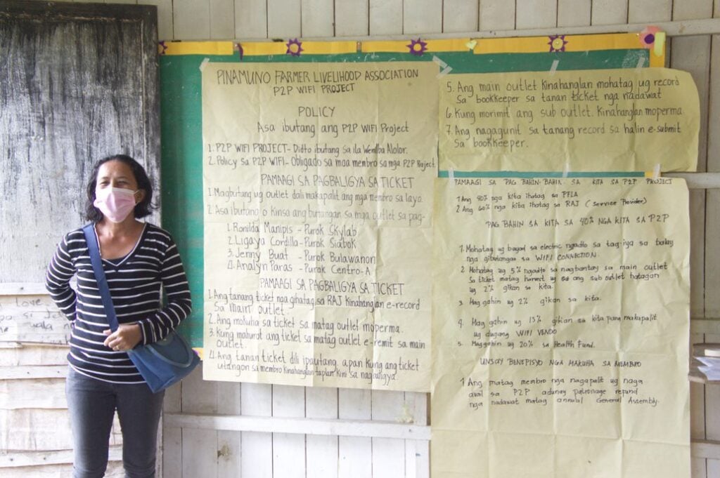 A member of the Pinamuno Farmer Livelihood Association standing next to some large pieces of paper on a wall. Written on the paper are policies for involvement in the P2P project.