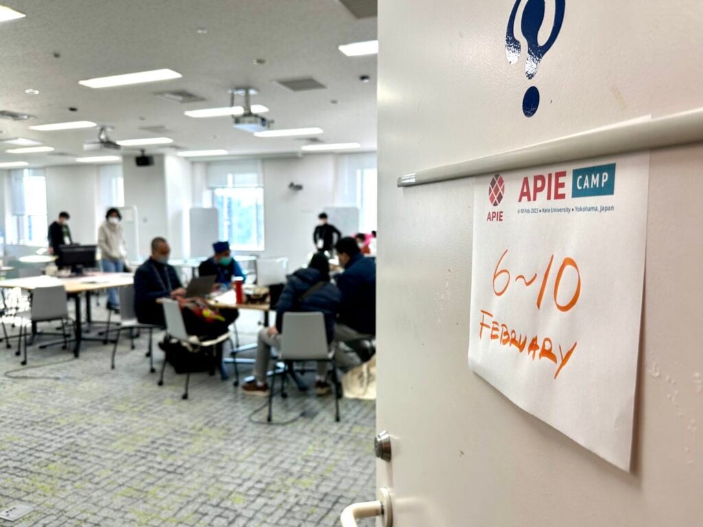 An image of an open door, leading to a room full of students working. The door has a sign on it with 6-10 February - the dates of the first APIE camp which was held in Japan.