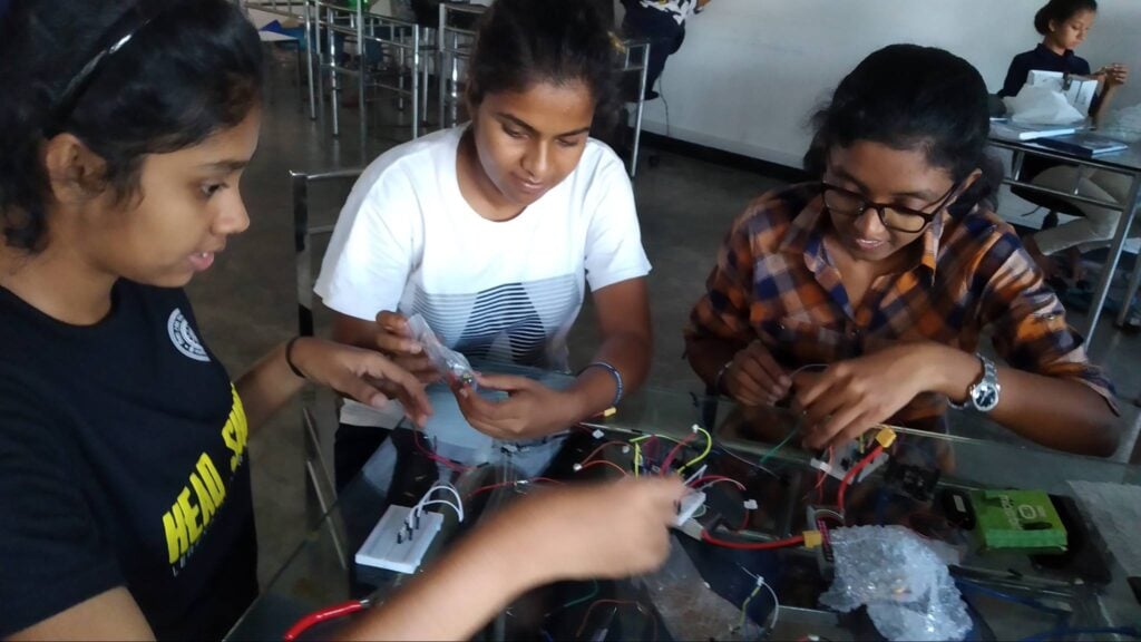 Three young women are shown working with wires and tech hardware on a table, while other young women work in the background.