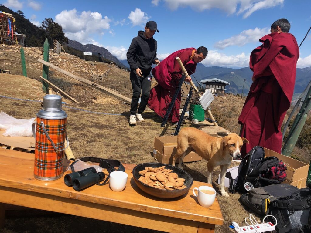 Picture shows two monks, a man in a black sweater, and a dog, as some equipment is being set up on a mountainside.