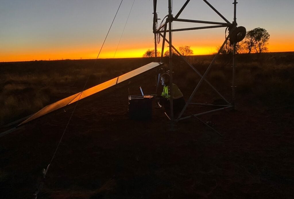 The picture shows a man working at a laptop underneath a tower frame and a solar panel, at sunset.