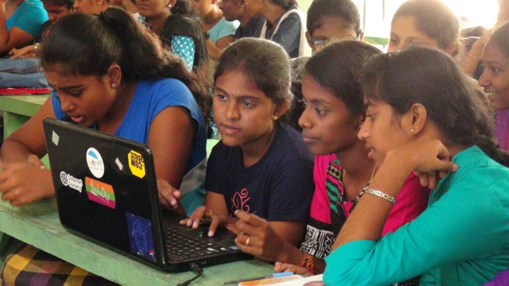 Image shows three girls working together on a laptop and another girl beside them.